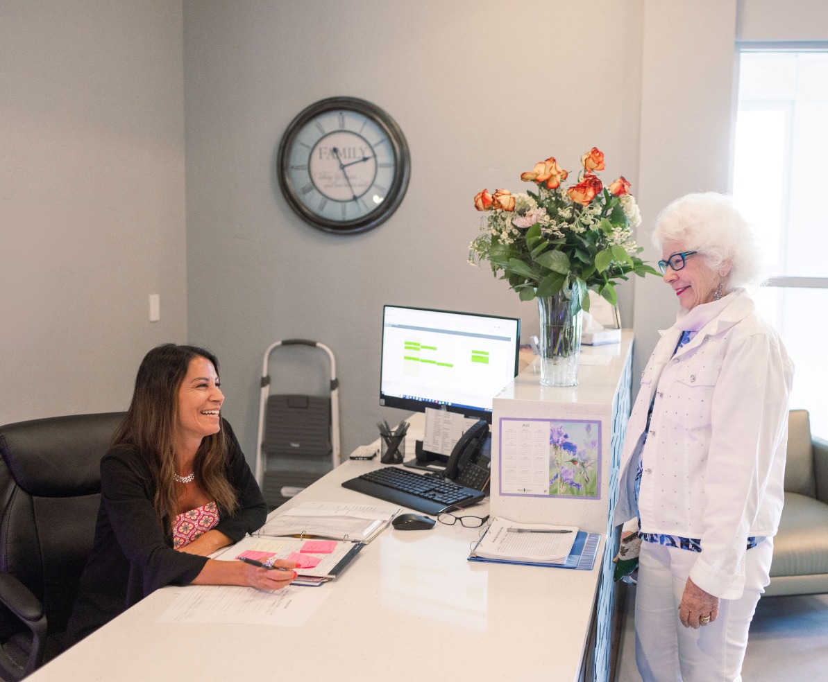 Senior woman stands at the reception desk, chatting with a staff member on the other side at The Summit of Coralville.
