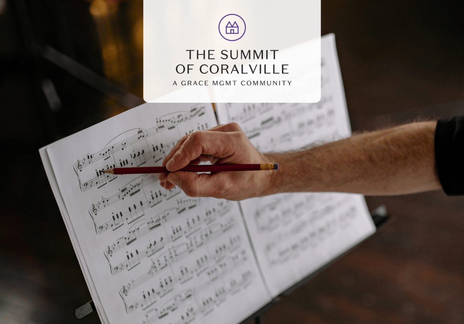 Hand holds a red pencil over an open sheet of music on a stand with a logo overlay reading 'The Summit of Coralville'